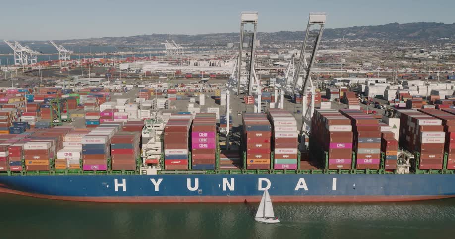 White sail boat floats beside a large cargo ship filled with shipping containers docked under cranes at a shipping yard
