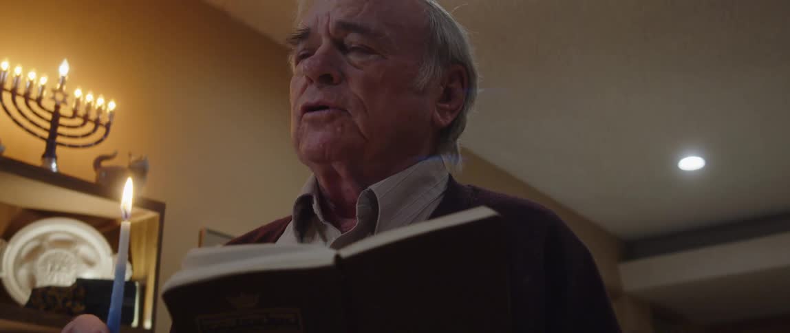 An old man talking holding a lit candle and a book while standing indoor at night