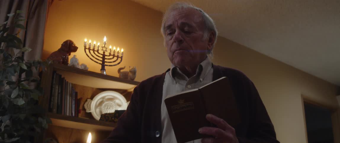 Elderly man reads from book while lighting a menorah