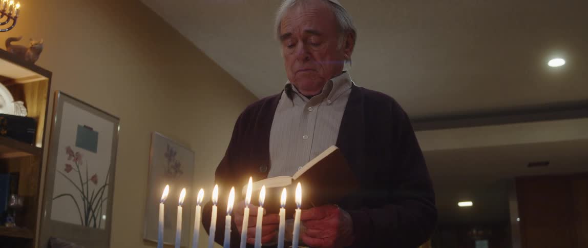 Elderly man at home stands in front of lit menorah and sings while reading from book