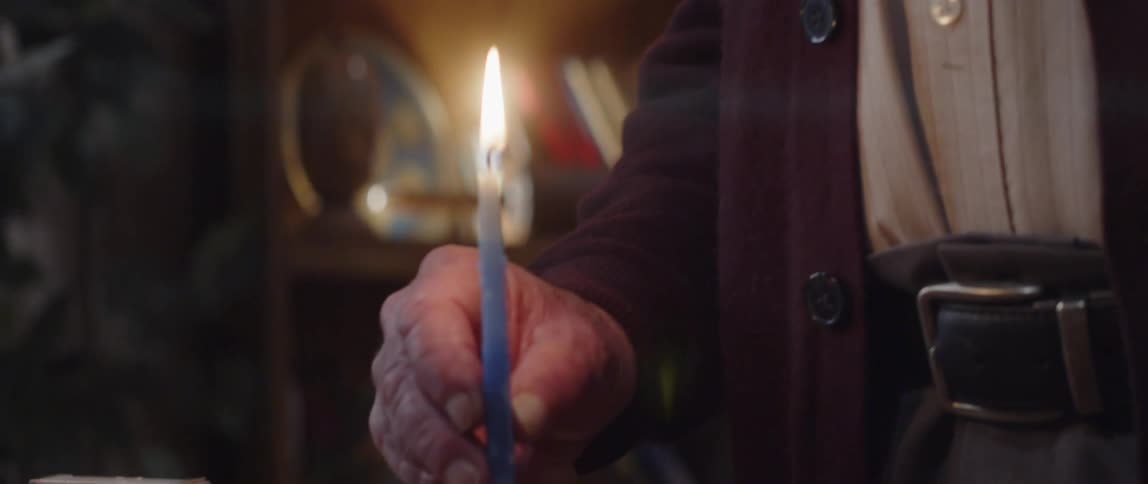 Elderly man standing indoor speaking while holding a lit candle at night