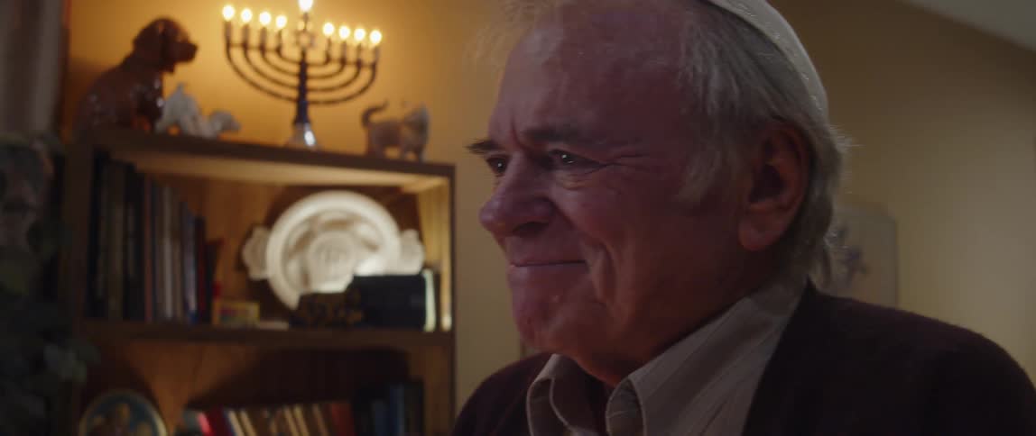 Elderly man wearing kippah smiles while standing at home with lit menorah in the background