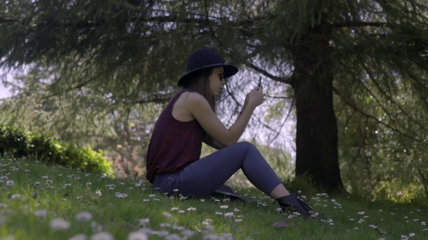 A woman sits on a grassy slope dotted with flowers and looks at her phone