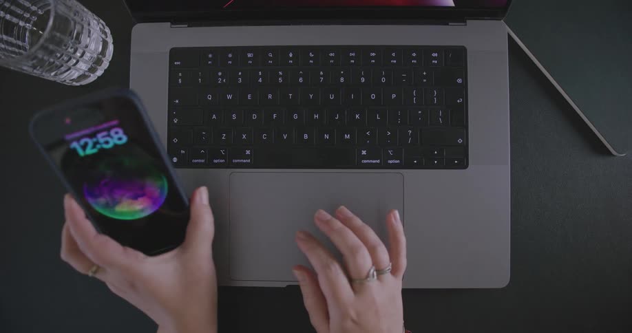 A phone sits next to a laptop computer on a table as a persons fingers are typing on the keyboard and stops quickly to look at the phone