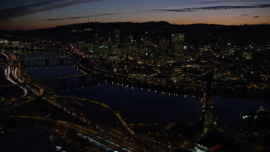 Aerial view of illuminated riverfront buildings and vehicles cruising on highways in the city