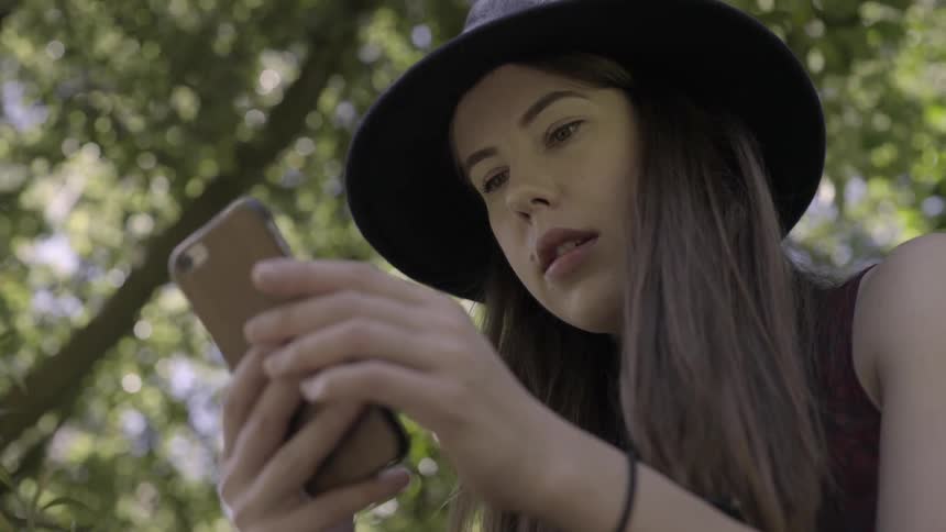Young lady wearing a hat using her phone while sitting under trees