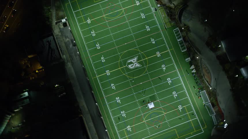 Football players move over brightly lit football field near busy highway at night