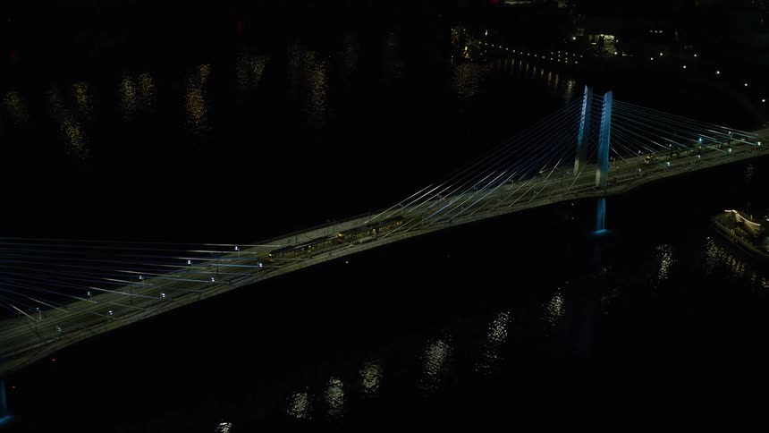 Two commuter trains pass over a suspension bridge above a river at night