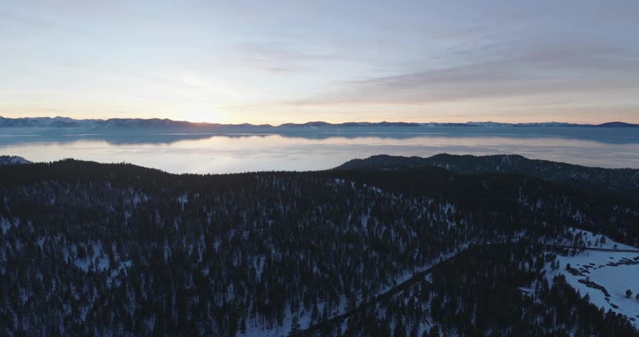 Aerial view of hills covered in pine trees and snow with a lake reflecting the white sky in the background with light from below the horizon