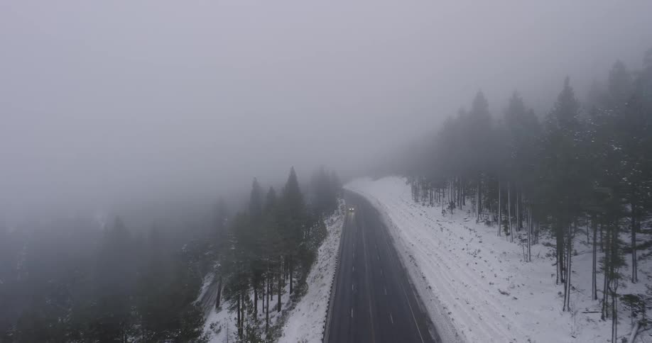 Light traffic moves on a snow covered forested mountain road during a blizzard