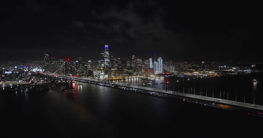 A huge suspension bridge over the water at night and behind it is a big city