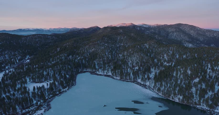 Aerial panorama of a mountain range amid conifers with snow under a pink dawn sky