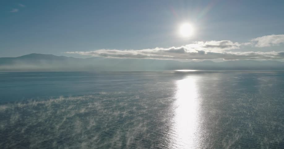 Sun hanging in a blue sky above white clouds reflecting across thin fog covered water below