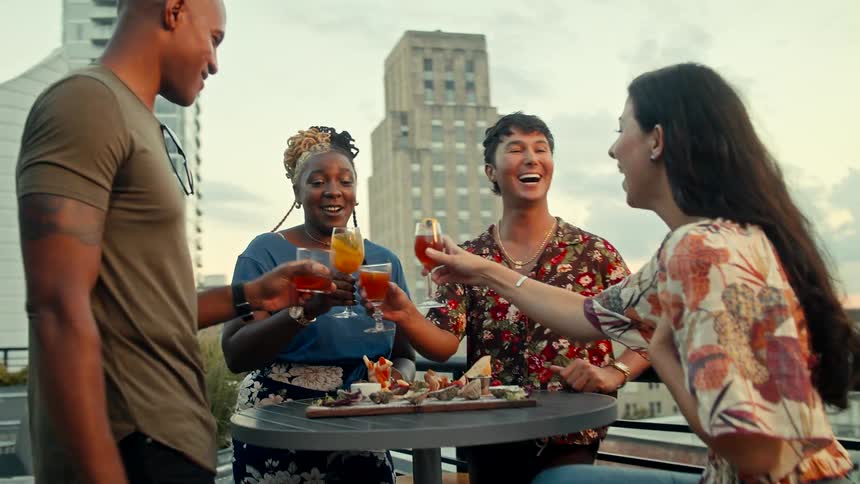 Three people sitting and one standing arrives and everyone toasts their glasses
