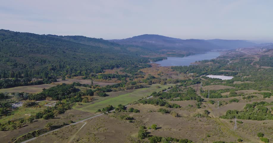 Aerial view of rural farmland and lake in a valley below a hill covered in lush green forest with mountains and hazy fog in the distance on a sunny day