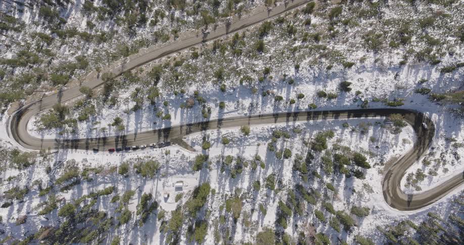 Aerial straight over a curvy mountain road surrounded by snow covered forests in the sunlight
