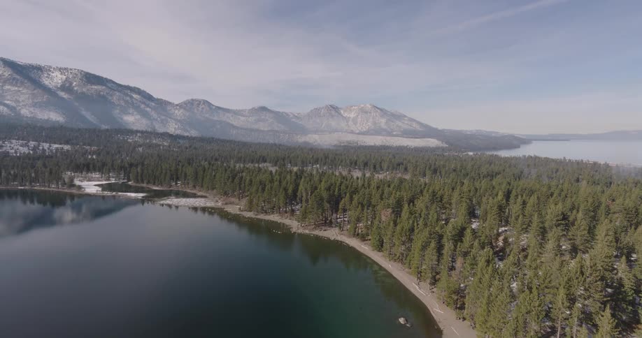 Aerial over a lake shore with dark reflecting water and pine trees with snow on the ground and mountain peaks under a hazy sky in the background