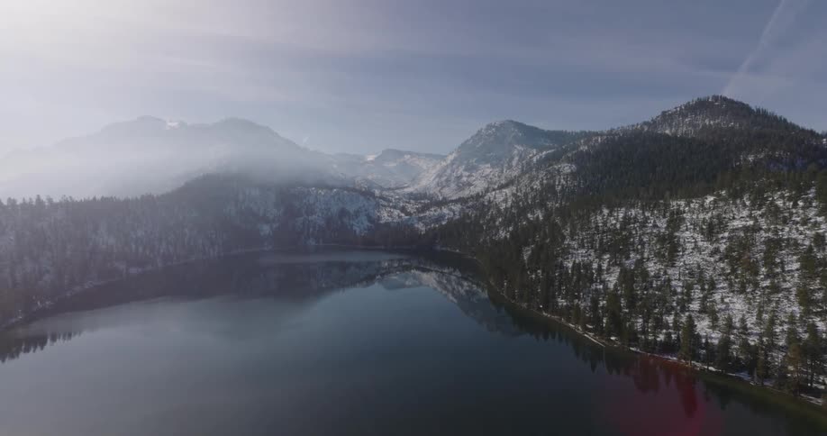 Aerial view of a lake and hillside with pine trees dusted in snow and fog covered mountains beyond the water