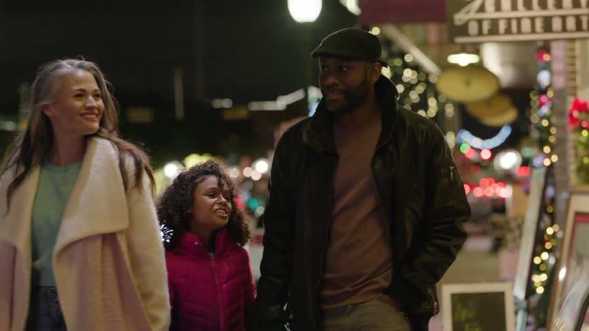 A family of a woman with a man and a child are walking on the sidewalk next to an establishment with several lights outside