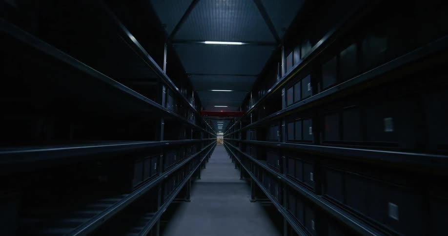 fpv drone down a corridor between two rows of floor to ceiling shelfs with black crate boxes on them and a door at the end