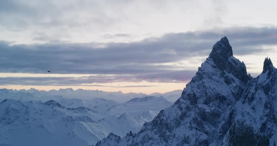 A bird flies near snow covered mountain peaks with mountains in the distance