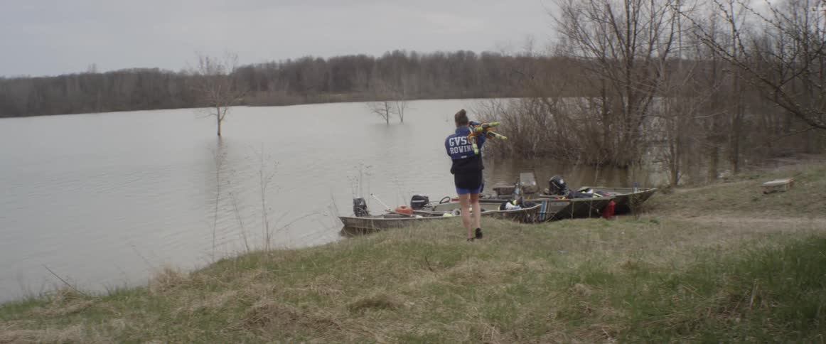 A man carries equipment toward two small boats on a lake shore