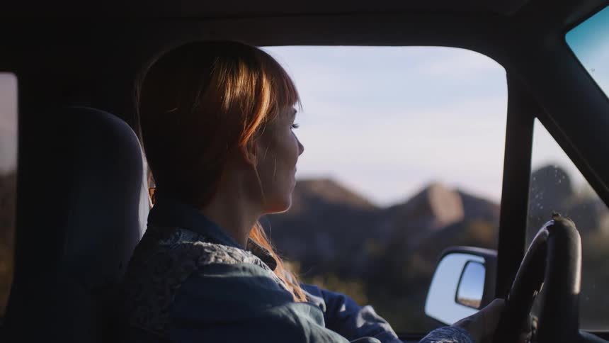 A woman is driving an automobile with mountains in the distance as she stops and rubs her neck
