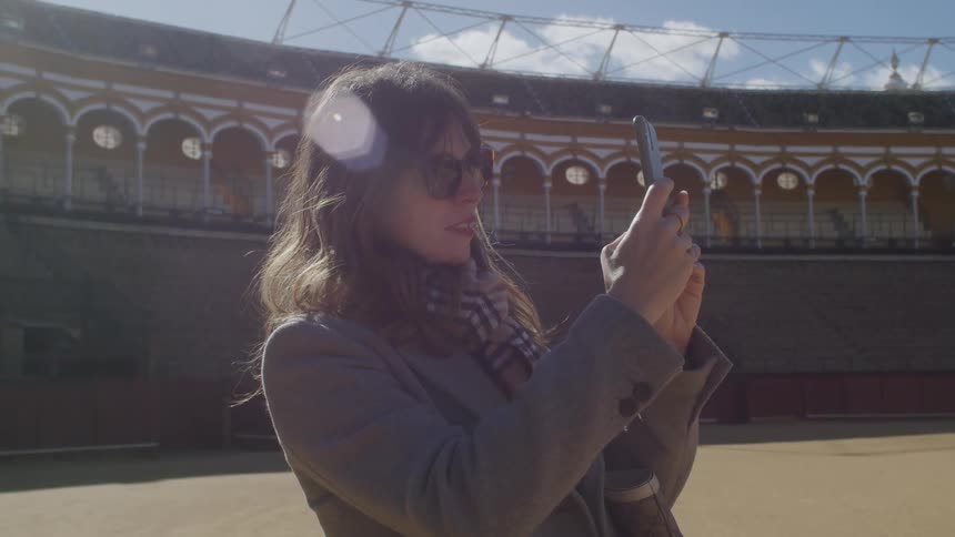 Woman stands in middle of historic open arena and takes a picture with her smartphone on a sunny day