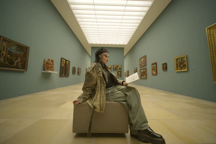 Woman in baggy clothes and heavy makeup sits reading in art museum