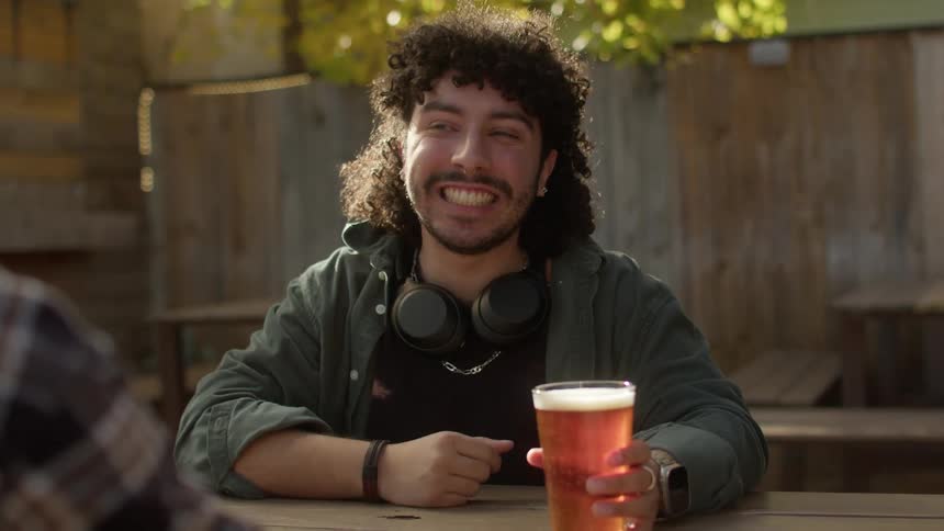 A man sitting is holding a glass of beer while talking and laughing with someone in front of him