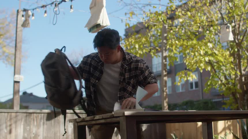 Man taking a book and notebook from his backpack and sitting at a wooden table in the backyard