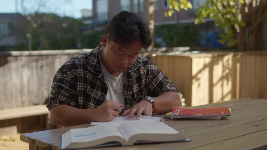A teenage boy is sitting outside at a table writing and doing his homework and he closes his notebook