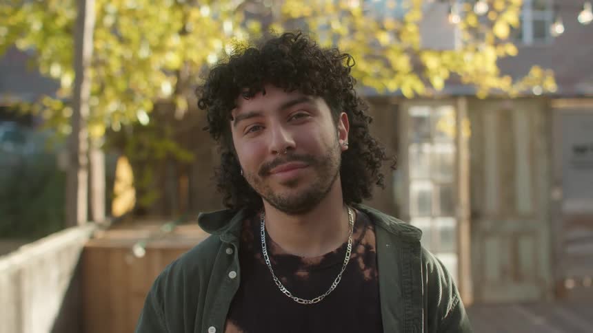 A man is standing outside with a fence in the background and he turns his head and smiles