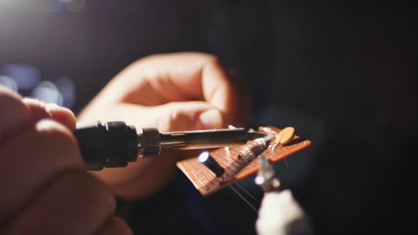 Closeup of a mans hands performing reballing on a hardware piece with the aid of a soldering tool emitting smoke