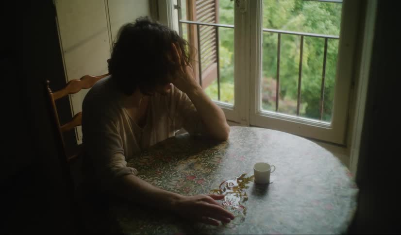 Man with long hair and a white t shirt sits at table by the windows