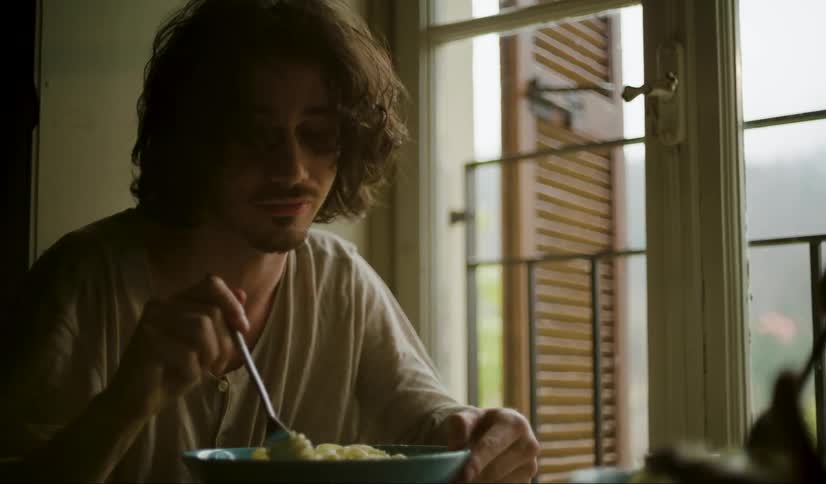 Man with long wavy hair and a white t shirt eats spaghetti sitting at the table