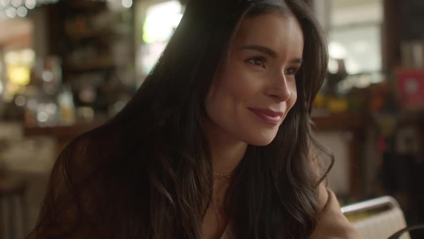 A woman sits at a table in a cafe holding a coffee mug and conversating with someone