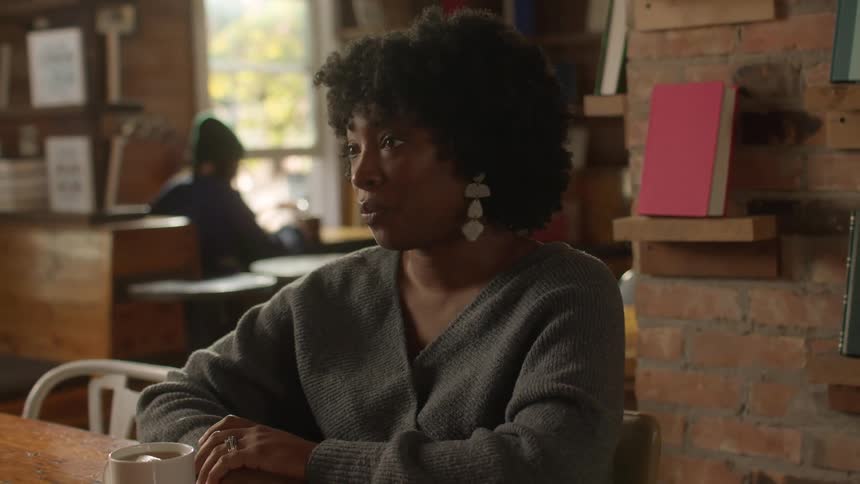 A black woman sits in a coffee shop while conversing with someone else