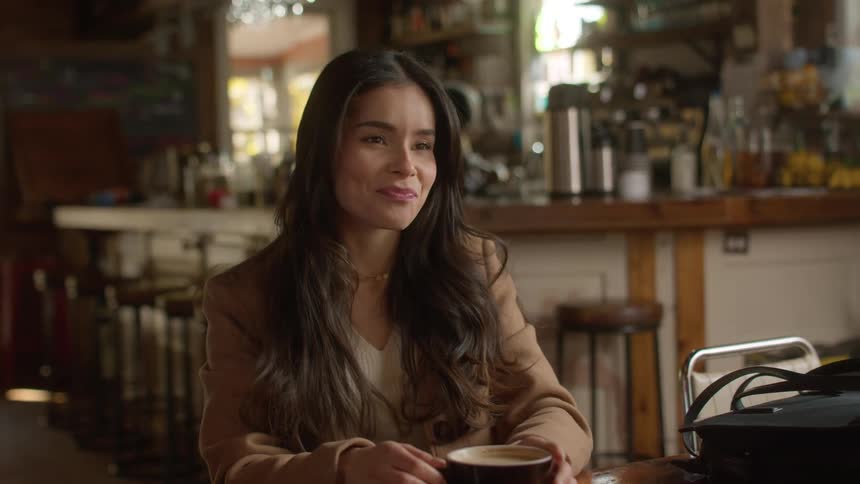 A woman with straight hair sporting a brown jacket sits at a table calmly sipping coffee from a black cup in the cafe
