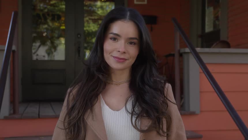 Woman with dark wavy hair a brown coat and white shirt smiles at camera in front of a cafe door