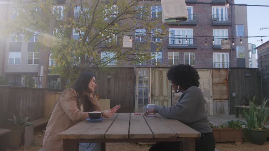 Two friends sit across from each other holding playing cards at a picnic table in back of a coffee shop where a worker hands off a coffee cup in the early evening before sundown