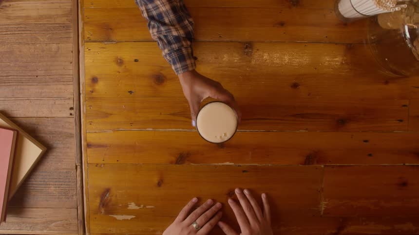 Closeup overhead view of one hand handing a glass of foamy drink to another on a wooden counter