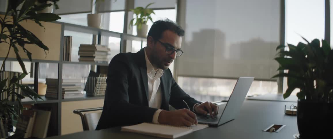 A man in formal clothes is in an office using a laptop on a table