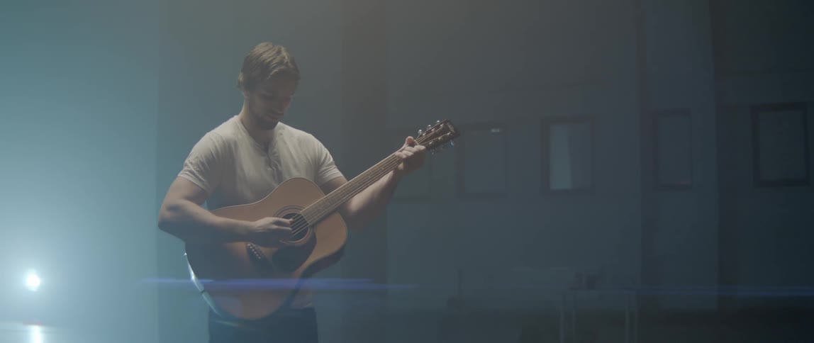 A man stands in a room alone playing an acoustic guitar