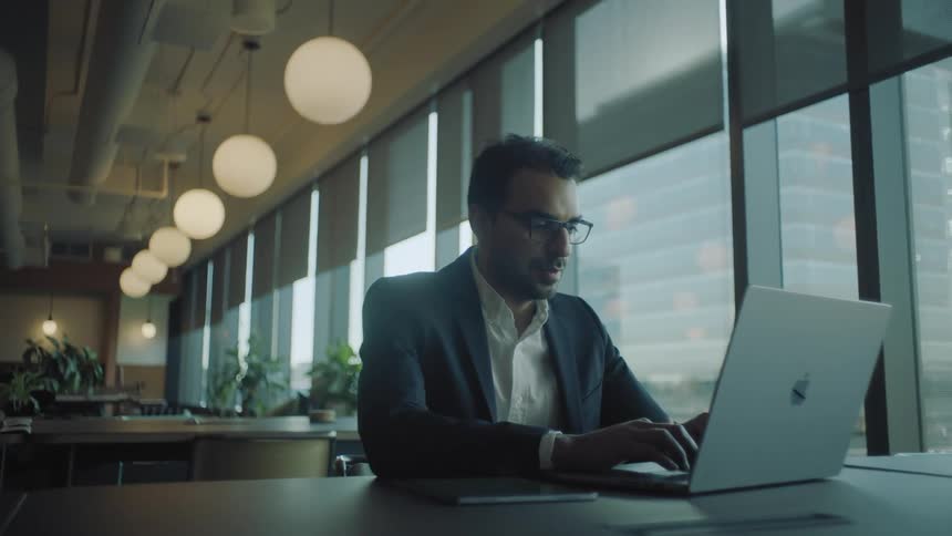 A man wearing glasses sits at a desk while typing on his laptop