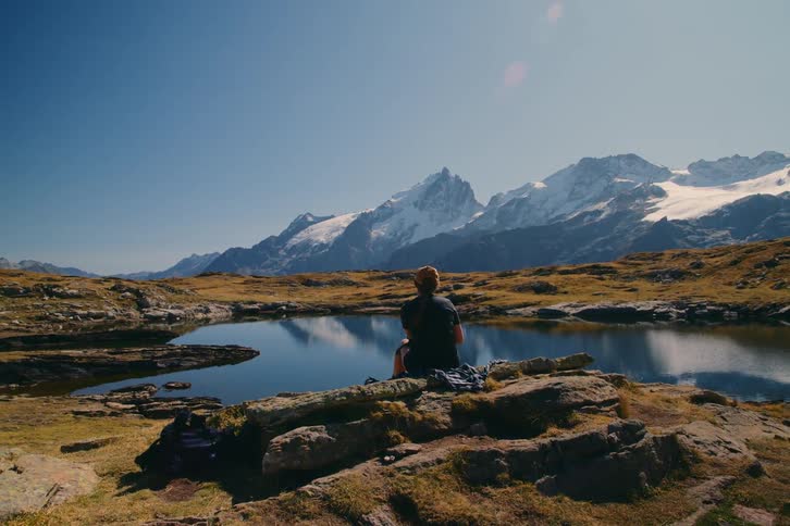 Woman sits on rocks beside a lake surrounded by highland and reflecting the mountains in the distance under a clear blue sky on a bright day