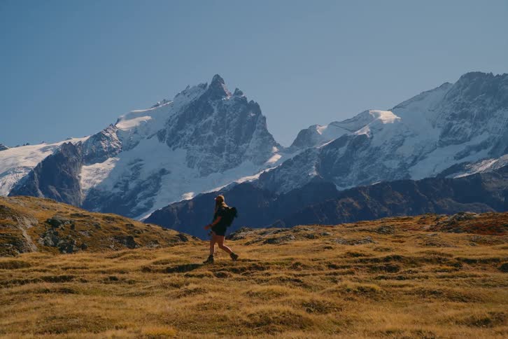 A hiker wearing a backpack walks along a mossy ridge with snowcovered mountains in the background on a clear sunny day