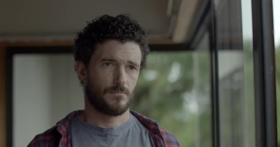 A man stands indoors looking out at the world with a serious emotional expression.