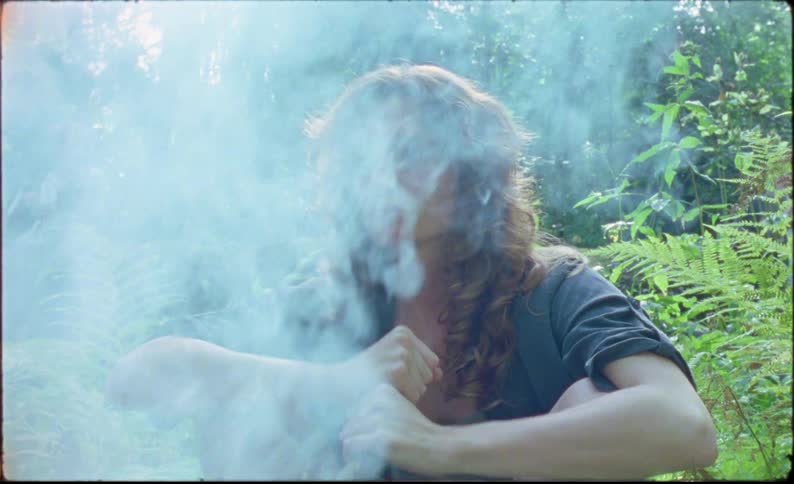 Young man with long hair sits near a campfire in the forest