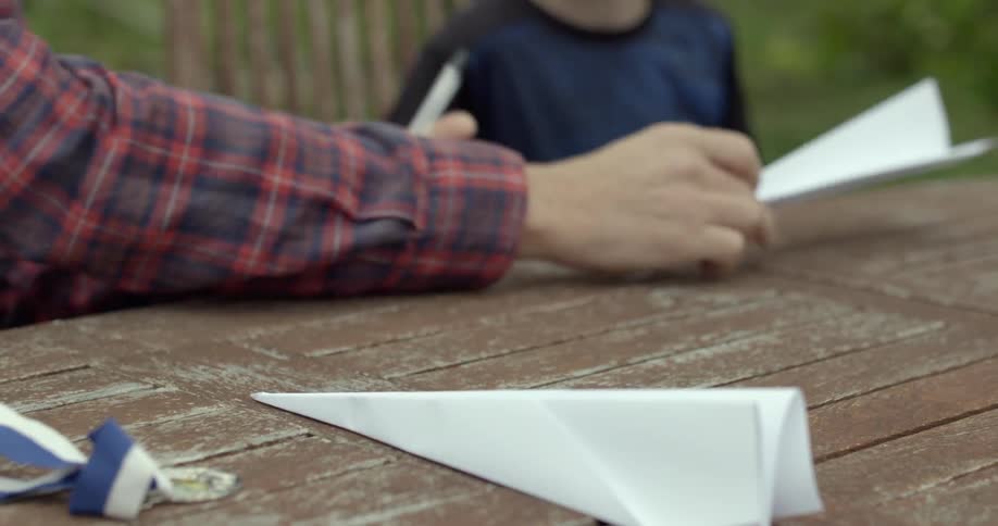 A man hands a pen and a paper airplane to his young son indoors.